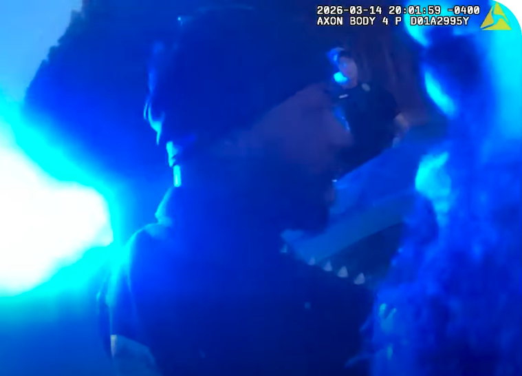 A close-up view of a person wearing a dark beanie and glasses, illuminated by intense blue light from the left side. The image appears to be from a body camera, as indicated by the timestamp and text at the top right corner. The background is dark, and the person's face is partially visible in profile.