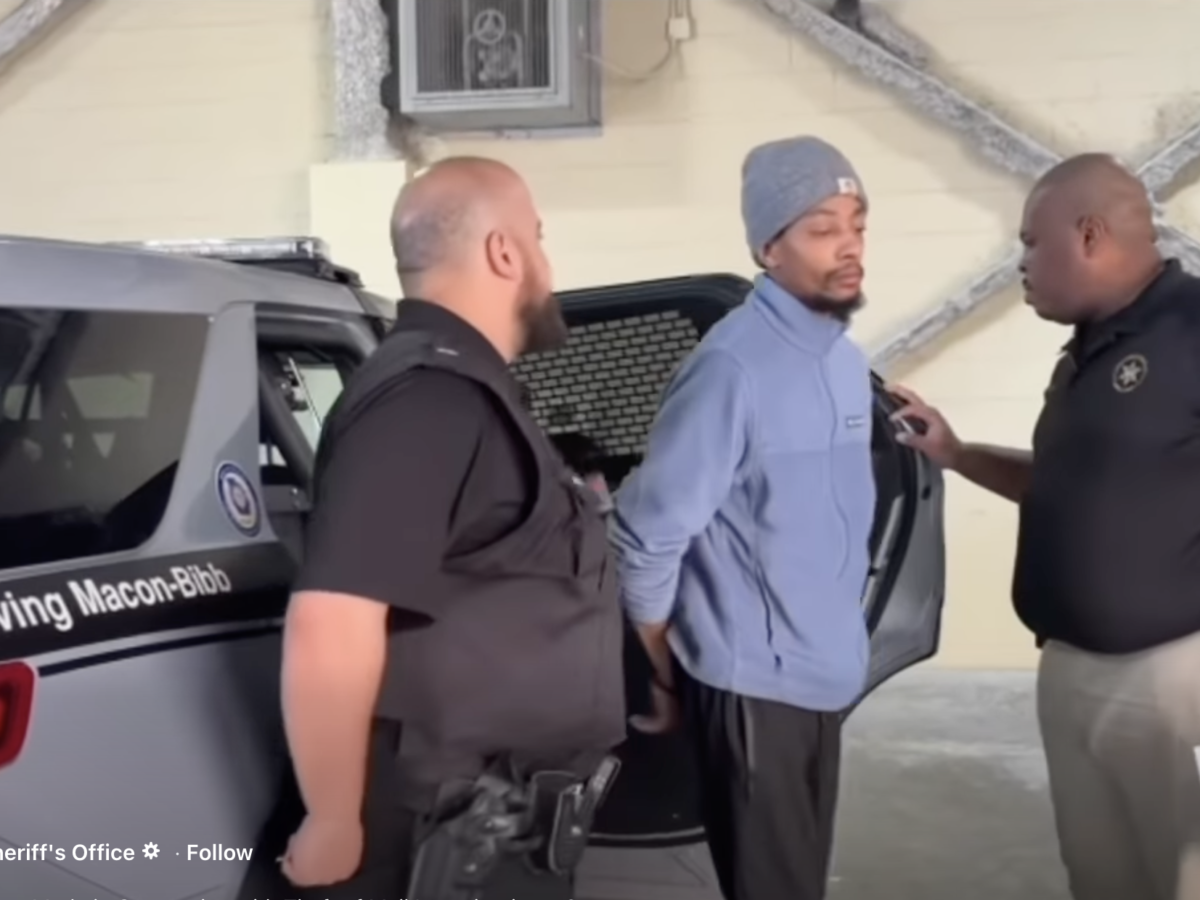 Two law enforcement officers are detaining a man in a light blue jacket and gray beanie. The man is standing with his hands behind his back near the open door of a police vehicle marked "Serving Macon-Bibb." One officer, wearing a black vest, stands close behind the man, while the other officer, in a black shirt and beige pants, is speaking to him and holding a document. The setting appears to be indoors with a beige wall and exposed structural beams in the background.