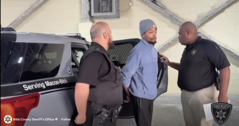Two law enforcement officers are detaining a man in a light blue jacket and gray beanie. The man is standing with his hands behind his back near the open door of a police vehicle marked "Serving Macon-Bibb." One officer, wearing a black vest, stands close behind the man, while the other officer, in a black shirt and beige pants, is speaking to him and holding a document. The setting appears to be indoors with a beige wall and exposed structural beams in the background.
