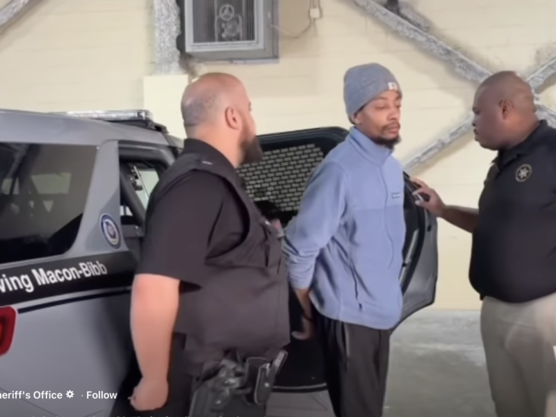 Two law enforcement officers are detaining a man in a light blue jacket and gray beanie. The man is standing with his hands behind his back near the open door of a police vehicle marked "Serving Macon-Bibb." One officer, wearing a black vest, stands close behind the man, while the other officer, in a black shirt and beige pants, is speaking to him and holding a document. The setting appears to be indoors with a beige wall and exposed structural beams in the background.