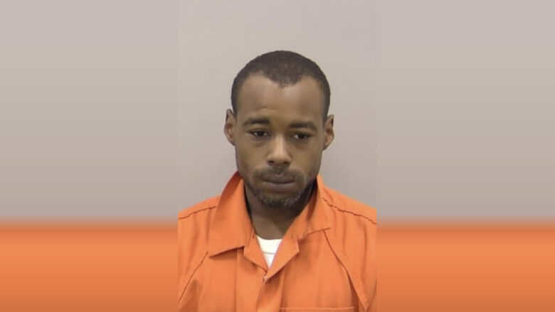 A man with short hair and a trimmed beard wearing an orange prison jumpsuit, looking slightly downward with a neutral expression.