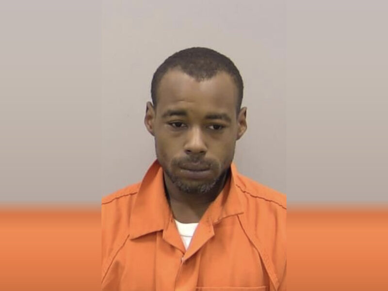 A man with short hair and a trimmed beard wearing an orange prison jumpsuit, looking slightly downward with a neutral expression.