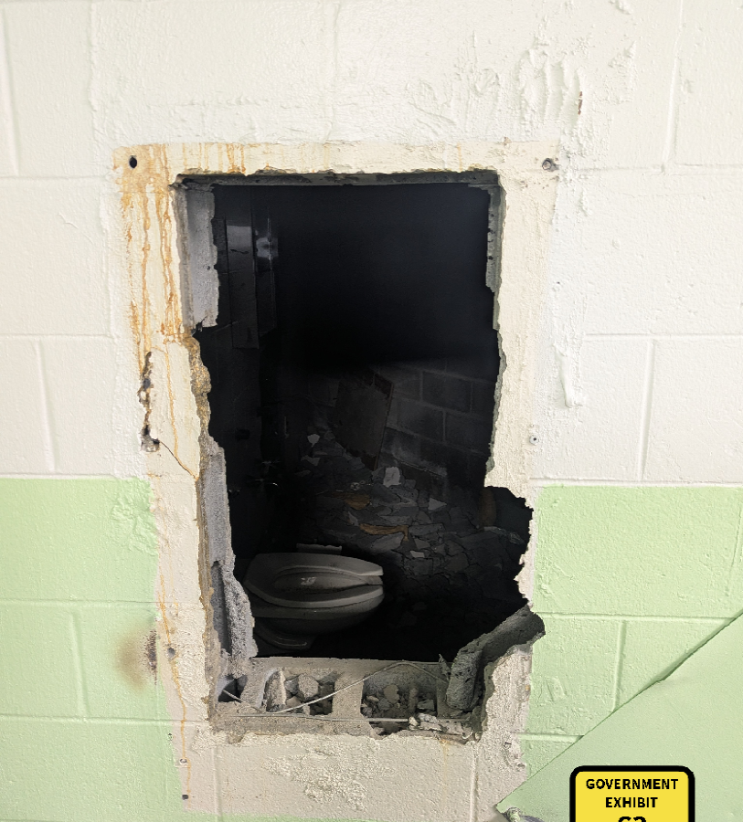 A damaged wall with a large rectangular hole revealing a dark, debris-filled space behind it. Inside the space, a broken toilet is visible. The wall is painted white on the upper half and light green on the lower half. Rust stains and chipped paint surround the hole. A yellow label in the bottom right corner reads "GOVERNMENT EXHIBIT 63.