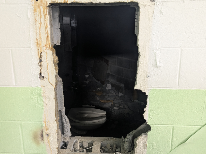 A damaged wall with a large rectangular hole revealing a dark, debris-filled space behind it. Inside the space, a broken toilet is visible. The wall is painted white on the upper half and light green on the lower half. Rust stains and chipped paint surround the hole. A yellow label in the bottom right corner reads "GOVERNMENT EXHIBIT 63.