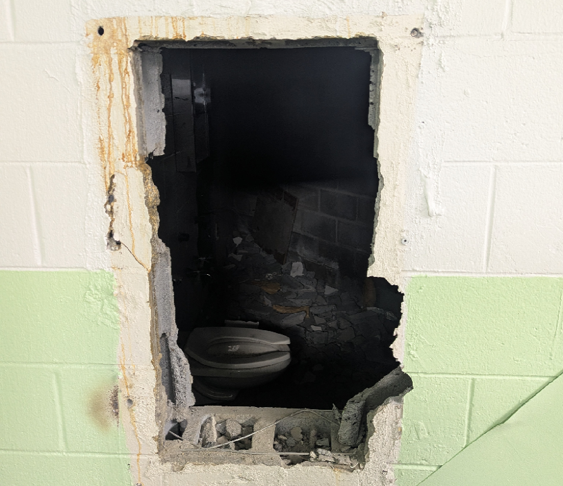 A damaged wall with a large rectangular hole revealing a dark, debris-filled space behind it. Inside the space, a broken toilet is visible. The wall is painted white on the upper half and light green on the lower half. Rust stains and chipped paint surround the hole. A yellow label in the bottom right corner reads "GOVERNMENT EXHIBIT 63.