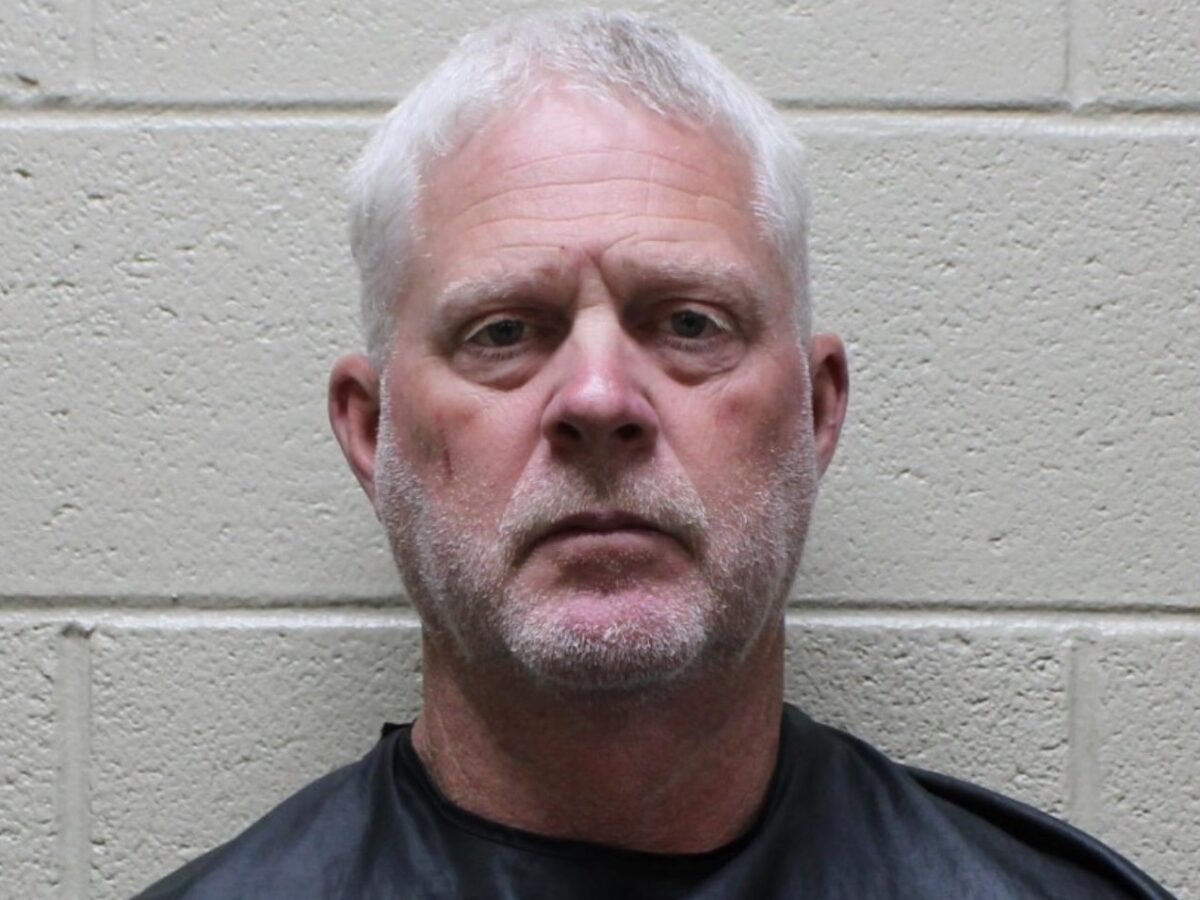 Middle-aged man with short white hair and a light beard, wearing a black garment, standing against a beige cinder block wall.