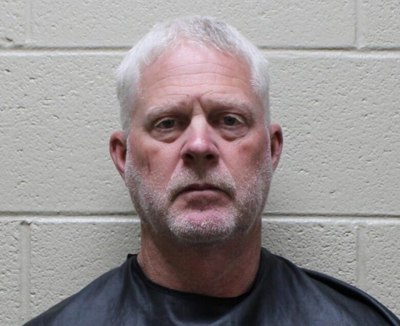 Middle-aged man with short white hair and a light beard, wearing a black garment, standing against a beige cinder block wall.