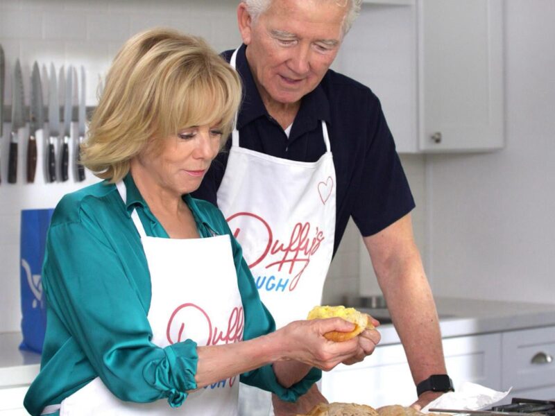 An older man and woman wearing white aprons with "Duffy's" written on them are in a kitchen. The woman, dressed in a teal blouse, is holding a piece of bread with a spread, while the man, in a black shirt, looks on. Various types of bread and a knife are on the counter in front of them. The kitchen has white cabinets and a knife rack on the wall.