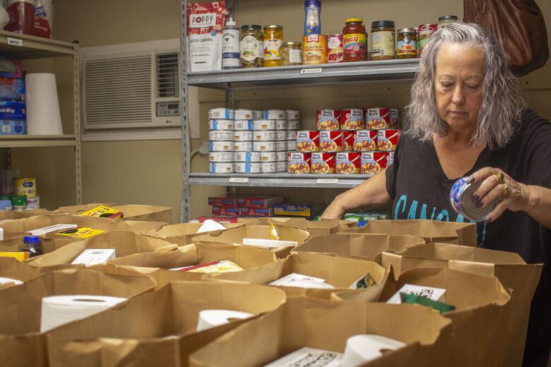 A woman with gray hair is sorting canned goods into multiple brown paper bags on a table. Behind her, metal shelves are stocked with various canned and jarred food items, including canned fruit and sauces. The setting appears to be a food pantry or storage room.
