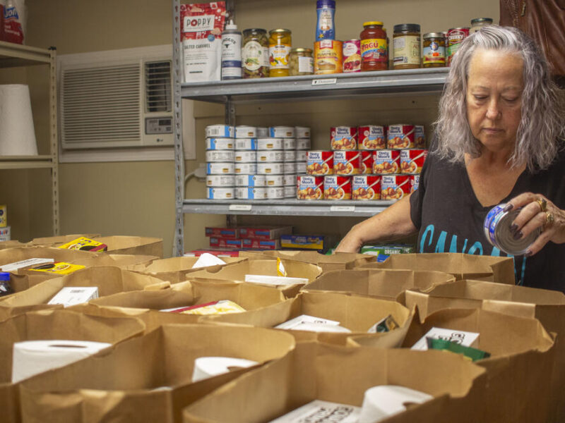 A woman with gray hair is sorting canned goods into multiple brown paper bags on a table. Behind her, metal shelves are stocked with various canned and jarred food items, including canned fruit and sauces. The setting appears to be a food pantry or storage room.