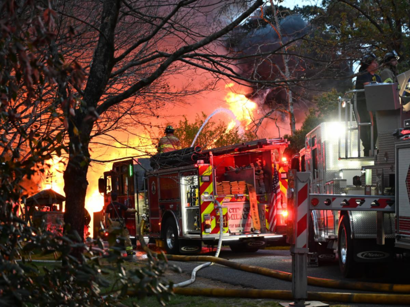 Firefighters are actively battling a large blaze with intense flames and thick black smoke visible in the background. Two fire trucks are present, with hoses connected and water being sprayed onto the fire. Firefighters in protective gear are seen working near the trucks and on top of one truck, surrounded by trees and vegetation. The scene is illuminated by the fire and emergency lights.