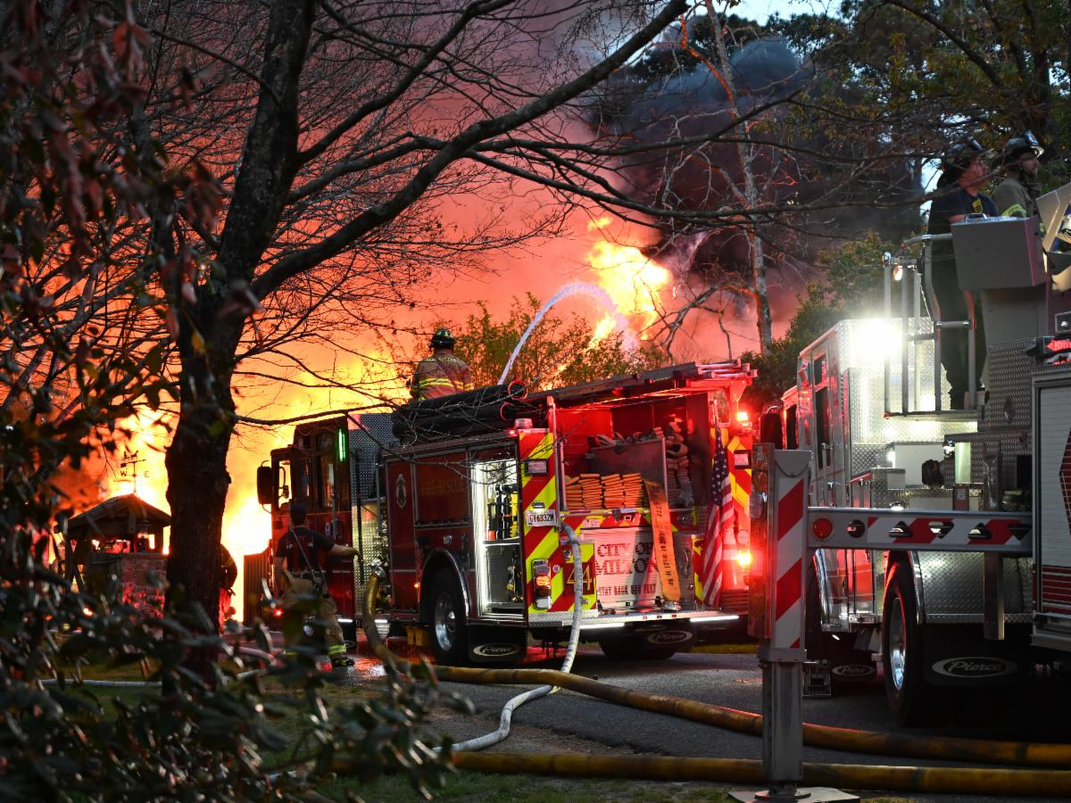 Firefighters are actively battling a large blaze with intense flames and thick black smoke visible in the background. Two fire trucks are present, with hoses connected and water being sprayed onto the fire. Firefighters in protective gear are seen working near the trucks and on top of one truck, surrounded by trees and vegetation. The scene is illuminated by the fire and emergency lights.