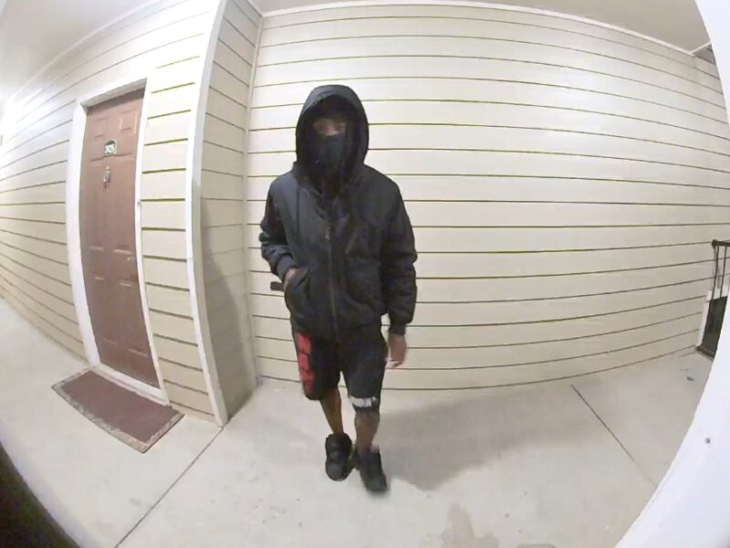 A person wearing a black hooded jacket with the hood up, a black face covering, black and red shorts, and black shoes is walking on a concrete floor in front of a beige wall with horizontal siding. There is a brown door with a white frame and a doormat to the left.