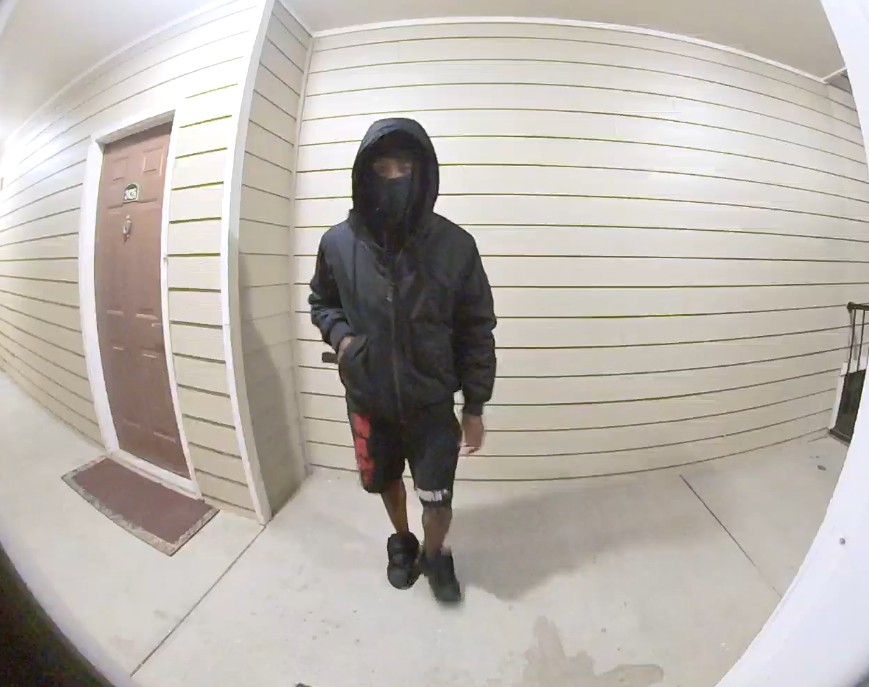 A person wearing a black hooded jacket with the hood up, a black face covering, black and red shorts, and black shoes is walking on a concrete floor in front of a beige wall with horizontal siding. There is a brown door with a white frame and a doormat to the left.