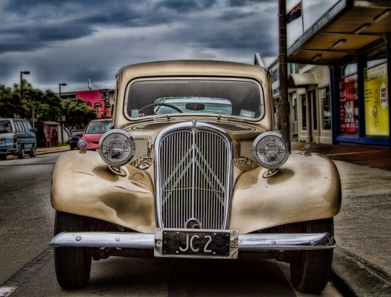 Front view of a vintage beige car with rounded fenders and a prominent chrome grille, parked on a street with other vehicles and buildings in the background under a cloudy sky. The license plate reads "JC2.