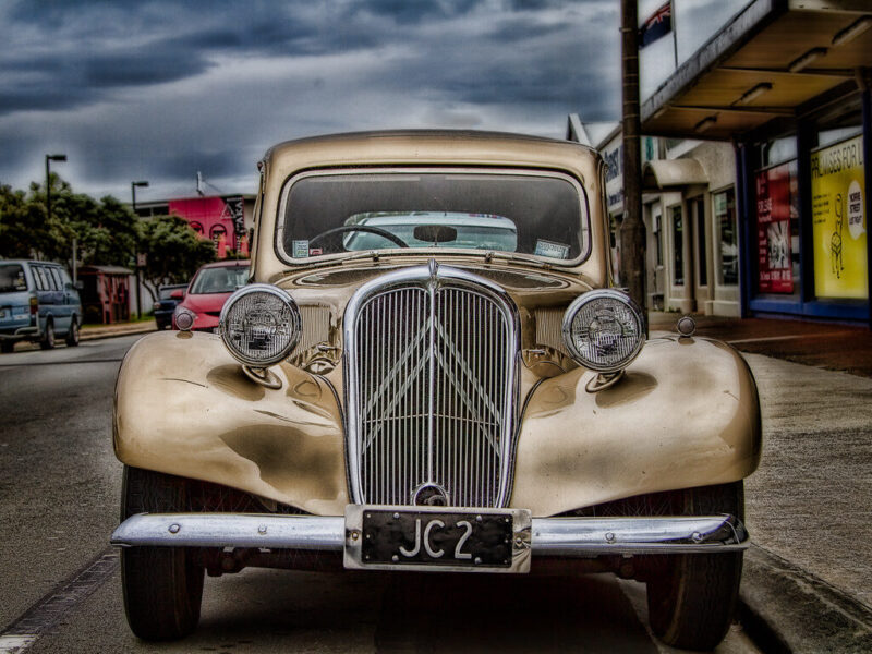 Front view of a vintage beige car with rounded fenders and a prominent chrome grille, parked on a street with other vehicles and buildings in the background under a cloudy sky. The license plate reads "JC2.