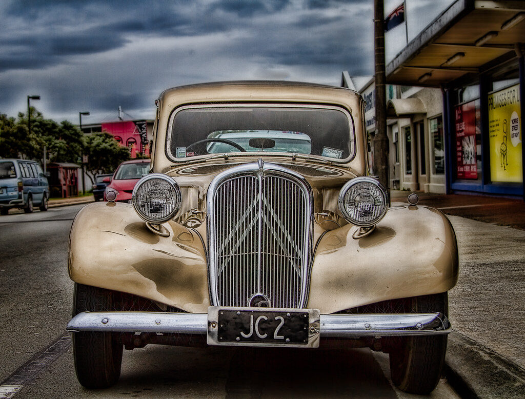 Front view of a vintage beige car with rounded fenders and a prominent chrome grille, parked on a street with other vehicles and buildings in the background under a cloudy sky. The license plate reads "JC2.