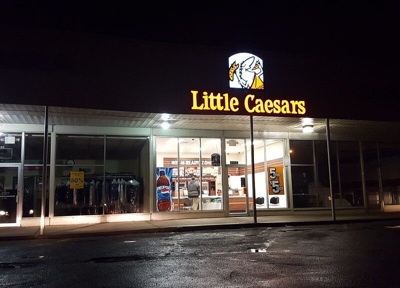 Little Caesars storefront at night with illuminated signage and interior lights. The entrance features glass doors and windows, with visible promotional signs inside, including a large Pepsi bottle image and a sign advertising "5 items $5 each." A person is standing inside near the counter. The surrounding area is dark with a wet pavement in front.