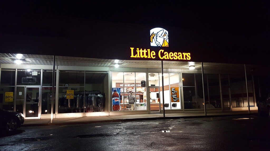 Little Caesars storefront at night with illuminated signage and interior lights. The entrance features glass doors and windows, with visible promotional signs inside, including a large Pepsi bottle image and a sign advertising "5 items $5 each." A person is standing inside near the counter. The surrounding area is dark with a wet pavement in front.