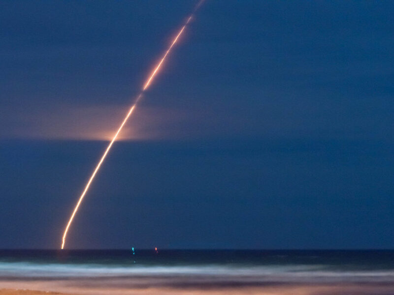 A bright, curved streak of light ascending into the night sky over a dark ocean, likely a long-exposure capture of a rocket launch or missile launch. The horizon shows a faint glow where the light originates, with the ocean below reflecting some light and gentle waves visible. The sky is mostly clear with a deep blue tone.