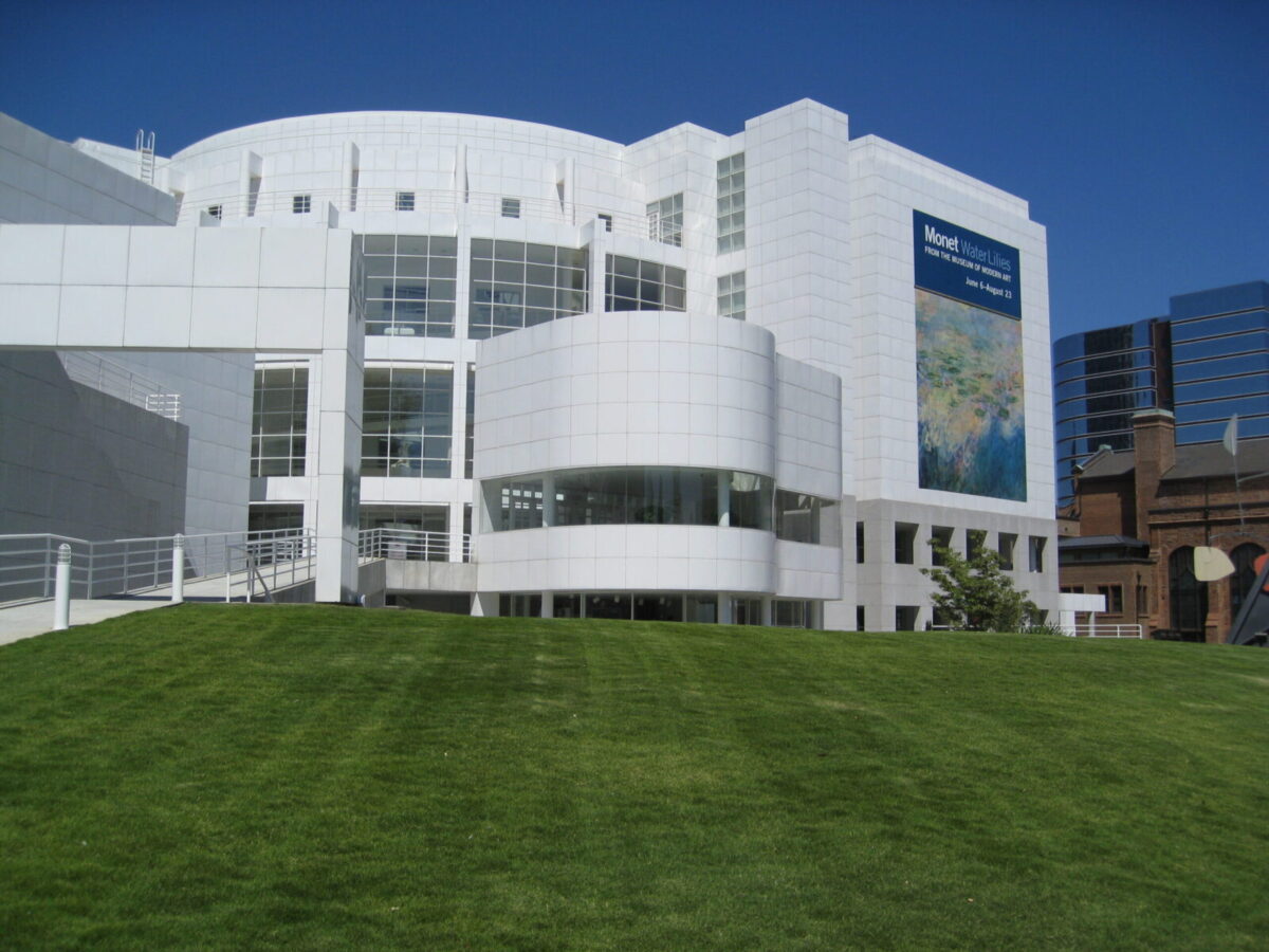 Modern white building with large windows and a curved section, situated behind a well-maintained green lawn. A large banner on the building advertises a Monet Water Lilies exhibition from the Museum of Modern Art, running from June 6 to August 23. The sky is clear and blue.