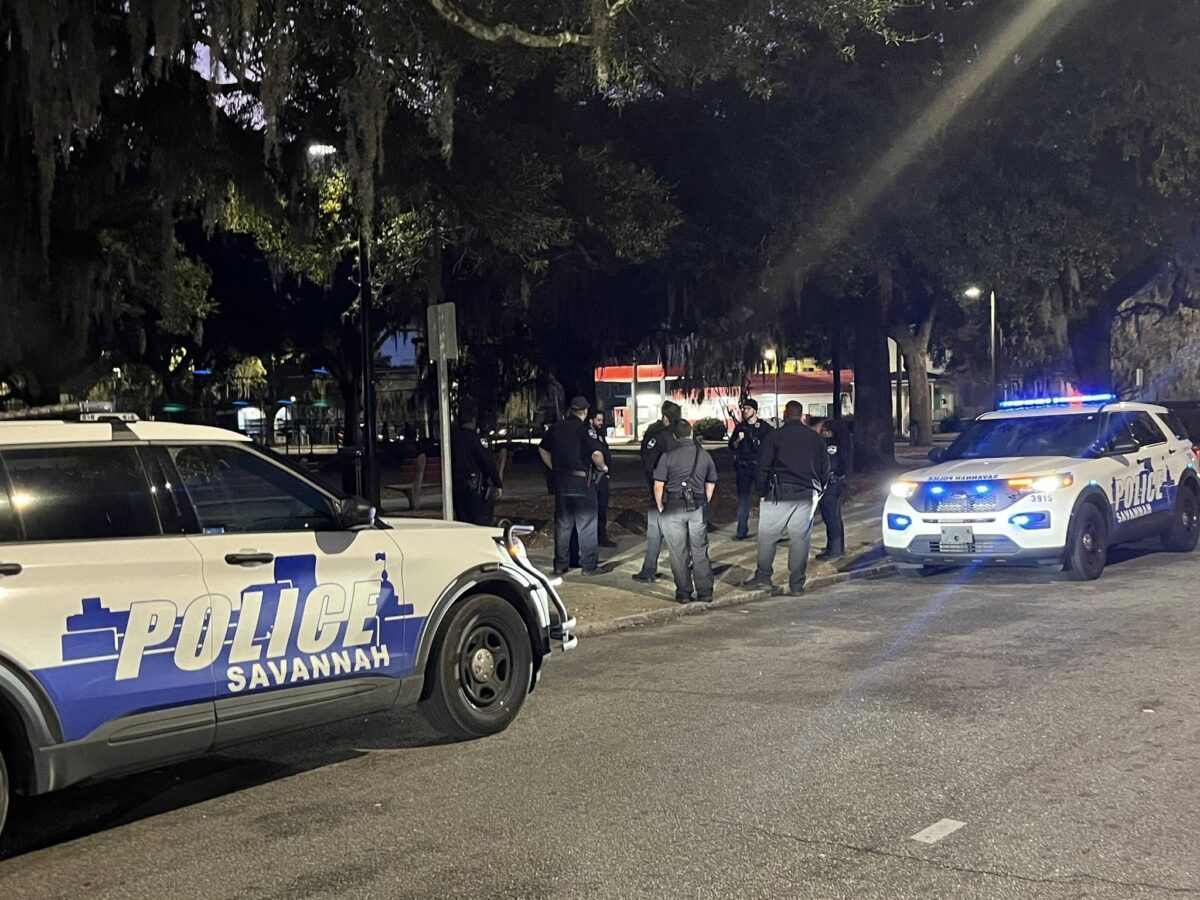 Two Savannah police SUVs are parked on a street at night with their lights on. Several police officers are standing on the sidewalk near the vehicles, engaged in conversation. The scene is dimly lit with trees and a building visible in the background.