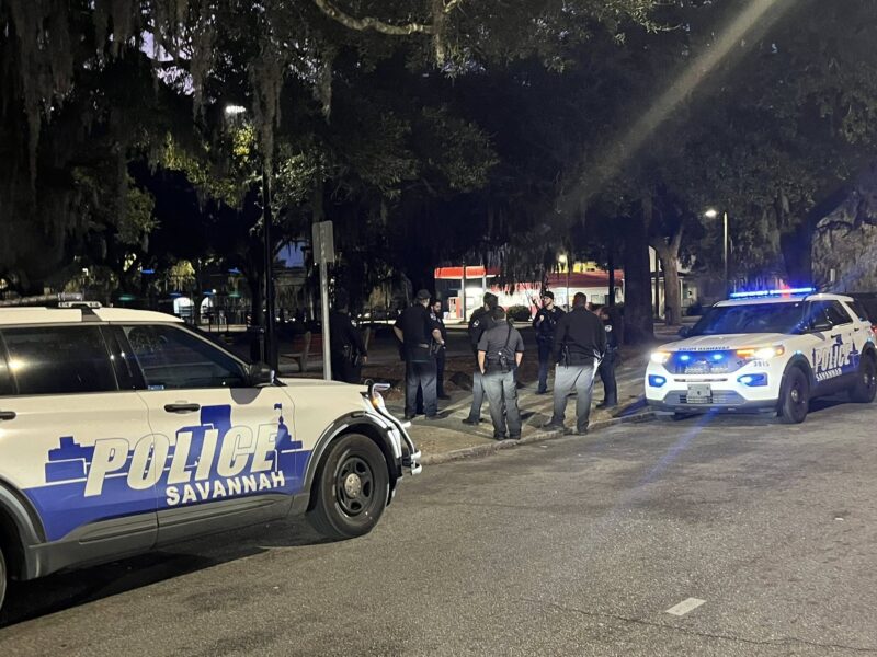 Two Savannah police SUVs are parked on a street at night with their lights on. Several police officers are standing on the sidewalk near the vehicles, engaged in conversation. The scene is dimly lit with trees and a building visible in the background.