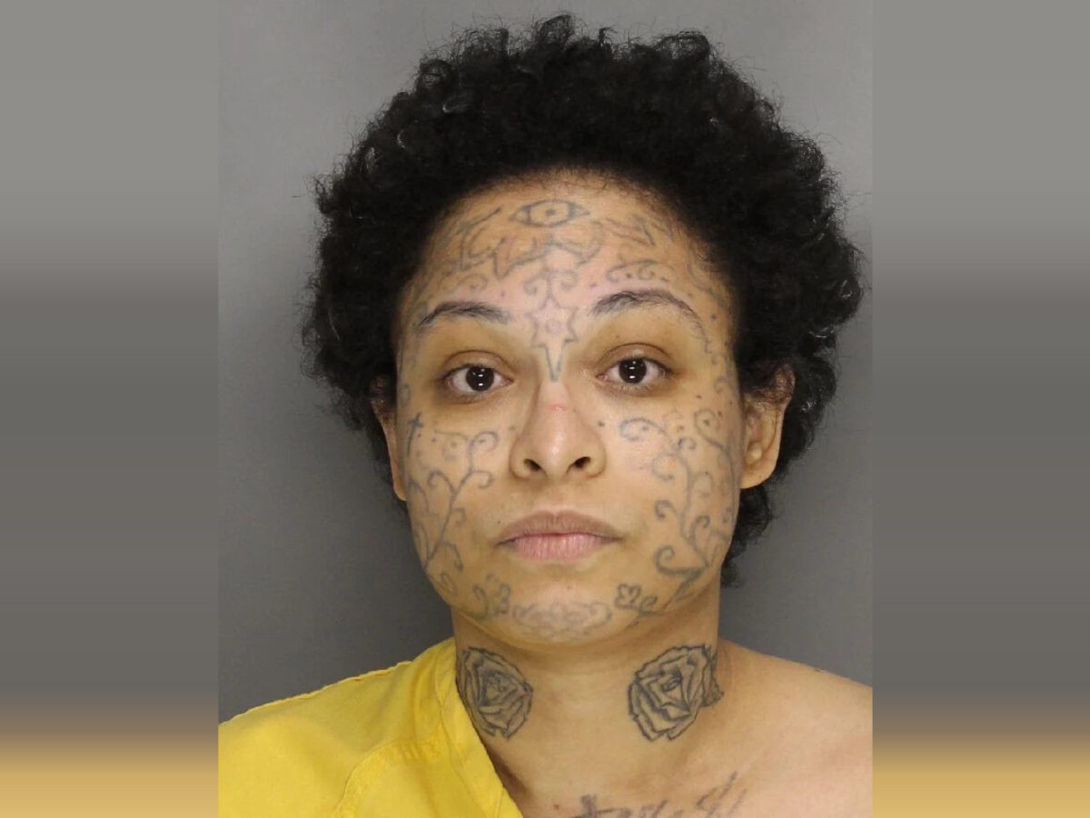 A person with short curly black hair and multiple tattoos on their face and neck. The facial tattoos include intricate swirling patterns and a prominent eye design on the forehead. The neck tattoos feature two roses. The person is wearing a yellow garment and has a neutral expression. The background is plain gray.