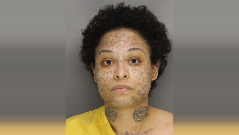 A person with short curly black hair and multiple tattoos on their face and neck. The facial tattoos include intricate swirling patterns and a prominent eye design on the forehead. The neck tattoos feature two roses. The person is wearing a yellow garment and has a neutral expression. The background is plain gray.