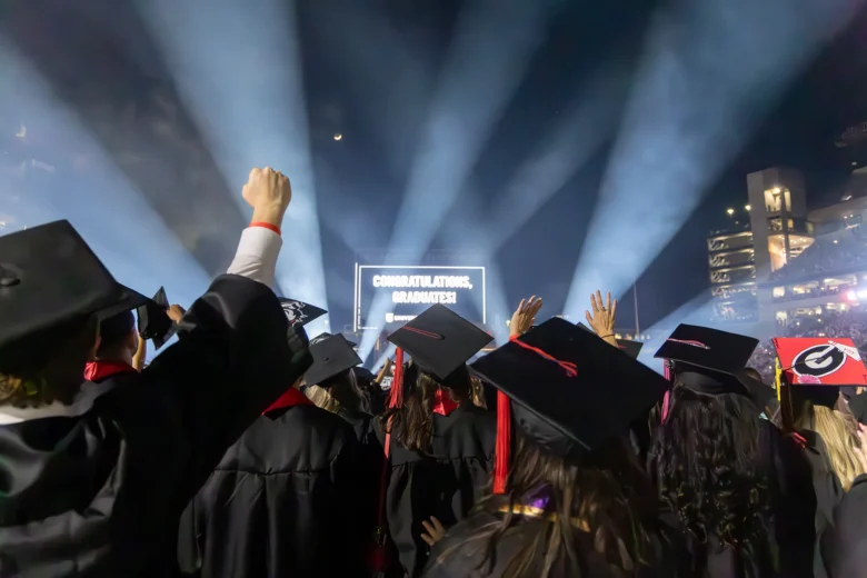 Graduates wearing black caps and gowns celebrate at a nighttime graduation ceremony with bright spotlights shining into the sky and a lit screen in the background displaying "CONGRATULATIONS, GRADUATES!" Some graduates have their hands raised in excitement.