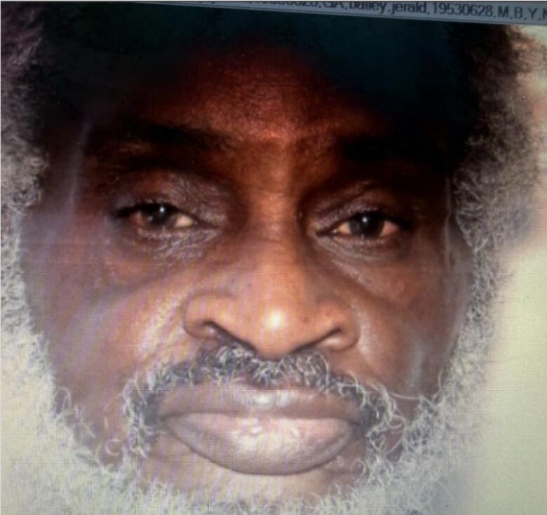 Close-up of an older man with dark skin, gray curly hair, and a full gray beard. His expression is neutral, and his eyes are looking directly at the camera.