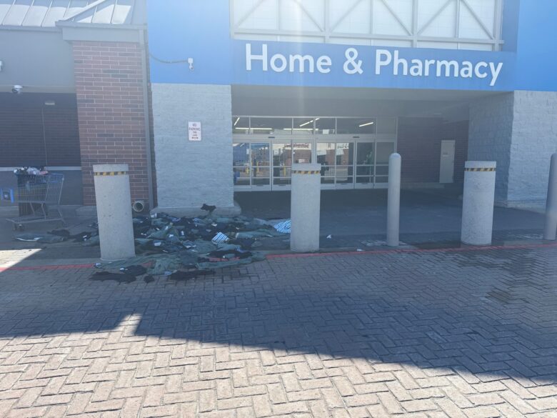 Entrance to a Home & Pharmacy store with a blue sign above the automatic sliding doors. In front of the entrance, there are three concrete bollards, and a large pile of dark-colored clothing or fabric scattered on the ground. A shopping cart filled with items is positioned to the left side near a brick wall. A "No Parking Fire Lane" sign is mounted on the wall near the entrance. The ground is paved with interlocking bricks.