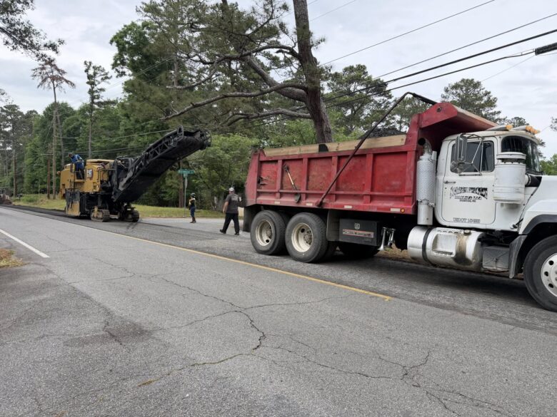 A large red and white dump truck is parked on the right side of a road, next to a yellow Caterpillar road milling machine. The milling machine has a conveyor belt extending towards the dump truck's bed, which is filled with asphalt material. Three workers are present: one standing on the milling machine, one walking on the road, and one standing near the truck. The scene is set on a tree-lined street with power lines overhead.