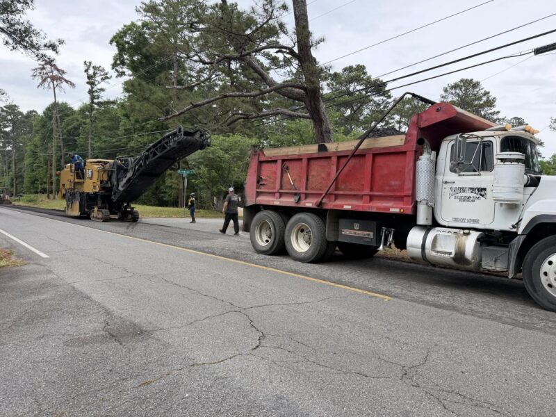 A large red and white dump truck is parked on the right side of a road, next to a yellow Caterpillar road milling machine. The milling machine has a conveyor belt extending towards the dump truck's bed, which is filled with asphalt material. Three workers are present: one standing on the milling machine, one walking on the road, and one standing near the truck. The scene is set on a tree-lined street with power lines overhead.