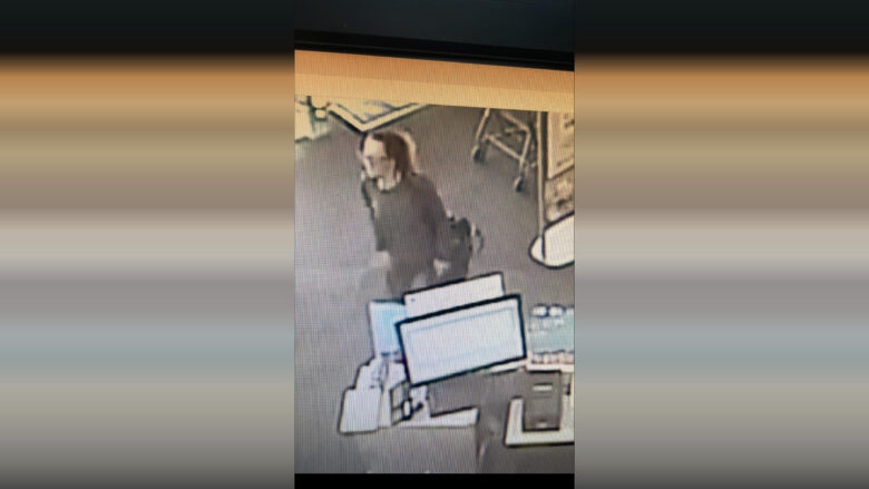 A person with long hair tied back, wearing a dark long-sleeve top, is walking inside a store near a checkout counter with computer monitors and a shopping cart in the background. The image appears to be from a security camera.