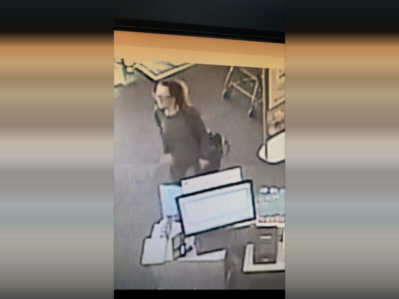 A person with long hair tied back, wearing a dark long-sleeve top, is walking inside a store near a checkout counter with computer monitors and a shopping cart in the background. The image appears to be from a security camera.