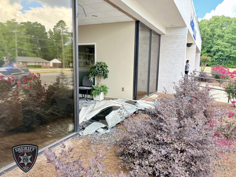 Broken glass and damaged window blinds lie on the ground outside a building with large glass windows. Inside, a chair and a potted plant are visible. The exterior features bushes with purple leaves and red flowers, and a person stands near the building's entrance. A sheriff's vehicle with flashing lights is reflected in the glass.