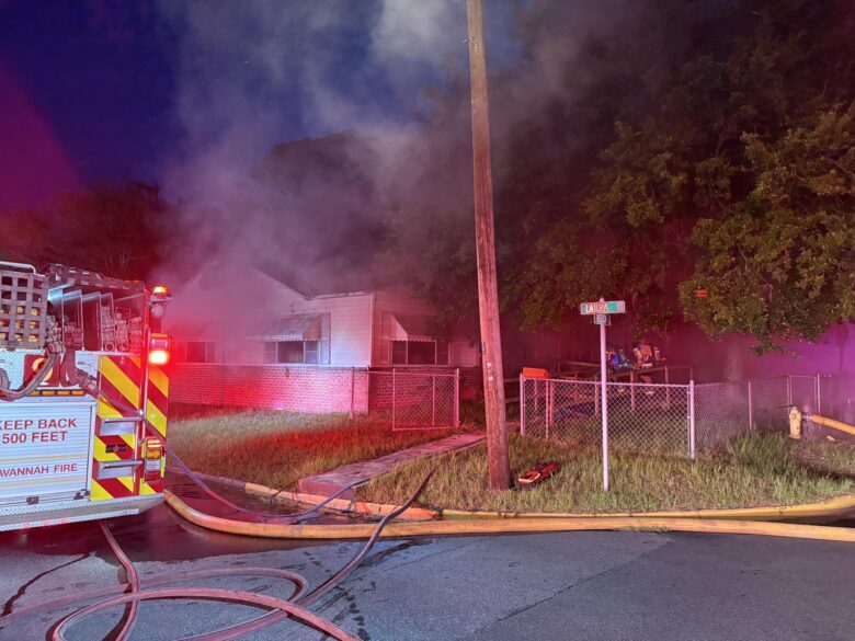 A nighttime scene shows a house with smoke billowing from it, indicating a fire. A fire truck from Savannah Fire is parked on the street with its lights flashing red. Fire hoses are laid out on the ground, leading towards the house. Two firefighters in gear are near the house, partially obscured by smoke and trees. A street sign reads "Laura Ave 100." The area is fenced, and a fire hydrant is visible on the right side of the image.