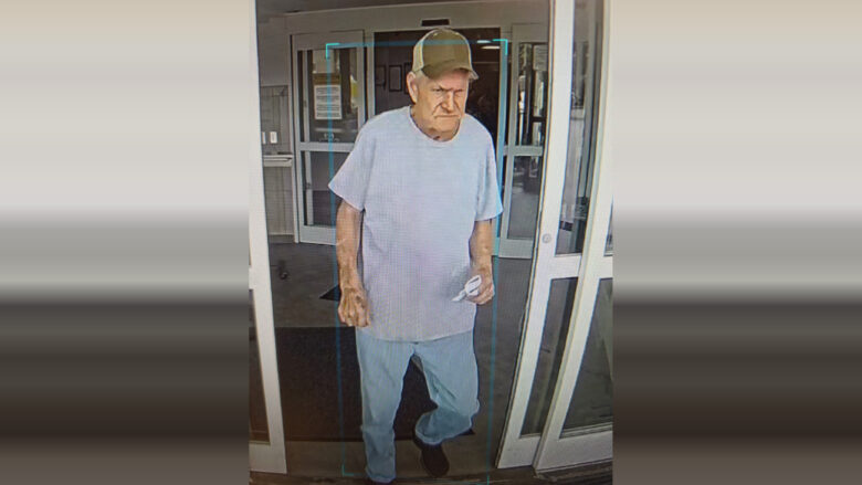 An elderly man wearing a light gray t-shirt, light blue jeans, black shoes, and a tan baseball cap is walking through a glass door. He is holding a small white object in his right hand. The background shows the entrance area of a building with glass doors and windows.