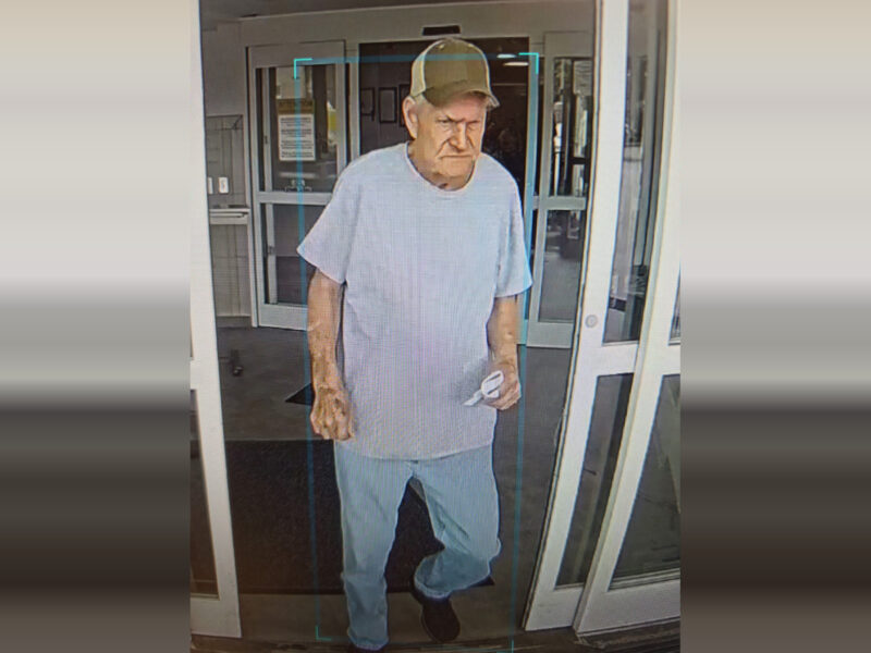An elderly man wearing a light gray t-shirt, light blue jeans, black shoes, and a tan baseball cap is walking through a glass door. He is holding a small white object in his right hand. The background shows the entrance area of a building with glass doors and windows.