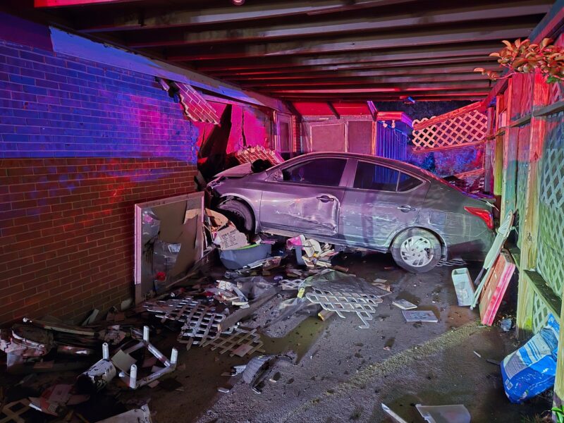 Silver sedan crashed into a brick wall under a wooden beam structure, causing significant damage to the car and the wall. Debris, including broken lattice panels, cardboard boxes, and various scattered items, covers the ground around the vehicle. The scene is illuminated by red and blue emergency lights.