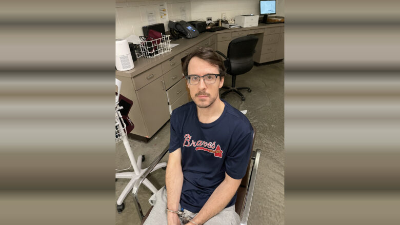 A man with glasses and short brown hair is seated in a chair with his hands handcuffed in front of him. He is wearing a navy blue "Braves" t-shirt and light gray pants. The background shows a room with beige cabinets, a black office chair, a computer monitor, a printer, and various office supplies on the countertop. The floor is concrete.