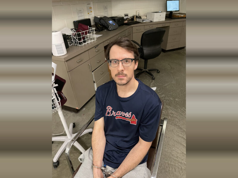A man with glasses and short brown hair is seated in a chair with his hands handcuffed in front of him. He is wearing a navy blue "Braves" t-shirt and light gray pants. The background shows a room with beige cabinets, a black office chair, a computer monitor, a printer, and various office supplies on the countertop. The floor is concrete.