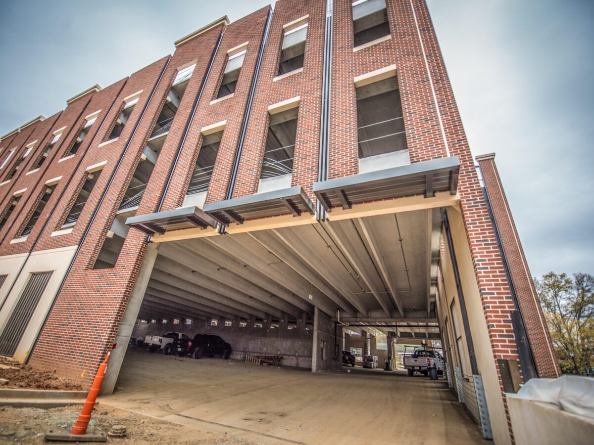 Multi-story brick parking garage with open window spaces and several vehicles parked inside, viewed from a low angle. The structure features concrete floors and ceilings with metal awnings above the entrance. An orange traffic cone is placed near the entrance on a dirt surface.