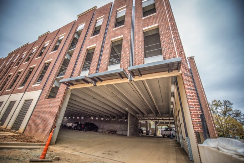 Multi-story brick parking garage with open window spaces and several vehicles parked inside, viewed from a low angle. The structure features concrete floors and ceilings with metal awnings above the entrance. An orange traffic cone is placed near the entrance on a dirt surface.
