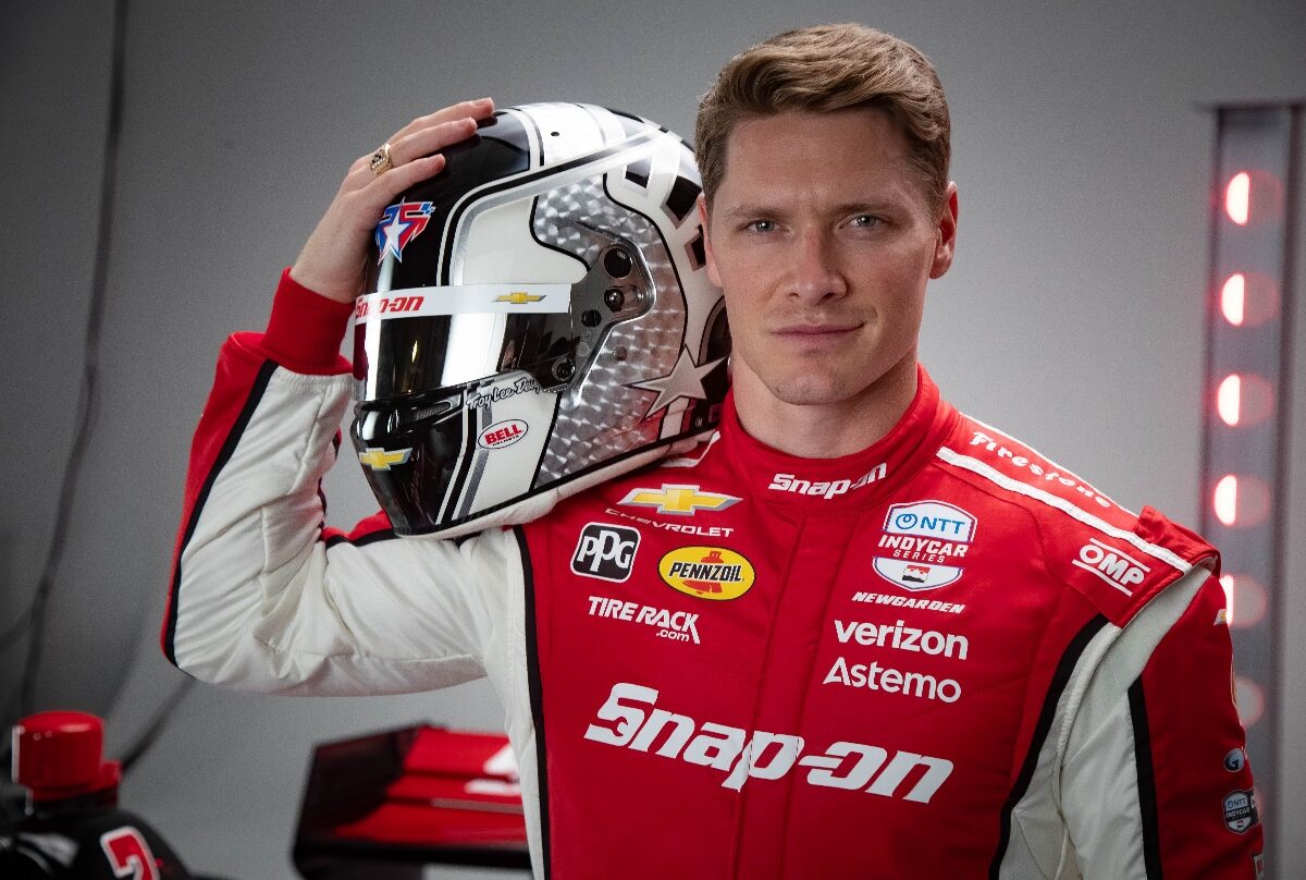 A man wearing a red and white racing suit with various sponsor logos, including Snap-on, Verizon, Pennzoil, and Chevrolet, holds a black, white, and silver racing helmet with a star design and multiple sponsor decals. The background is gray with red lights on the right side.