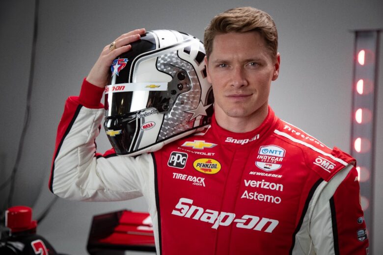 A man wearing a red and white racing suit with various sponsor logos, including Snap-on, Verizon, Pennzoil, and Chevrolet, holds a black, white, and silver racing helmet with a star design and multiple sponsor decals. The background is gray with red lights on the right side.