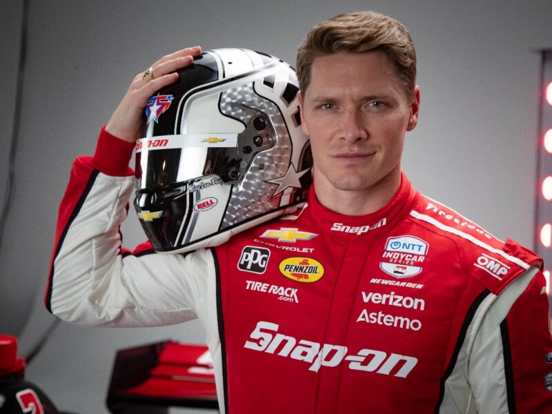 A man wearing a red and white racing suit with various sponsor logos, including Snap-on, Verizon, Pennzoil, and Chevrolet, holds a black, white, and silver racing helmet with a star design and multiple sponsor decals. The background is gray with red lights on the right side.