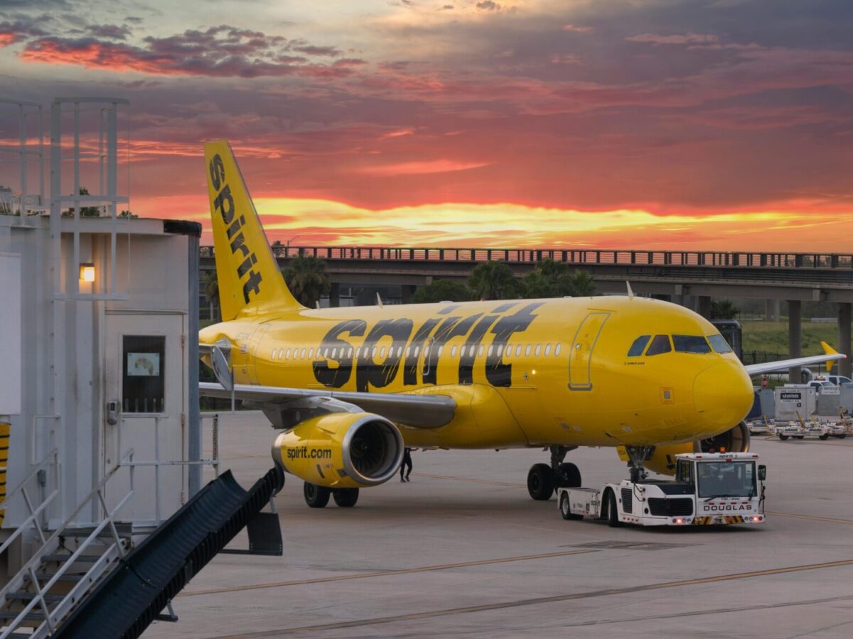 Yellow Spirit Airlines airplane parked at the gate during sunset, with a jet bridge on the left and a tug vehicle in front of the plane on the tarmac. The sky is filled with vibrant orange and purple clouds.