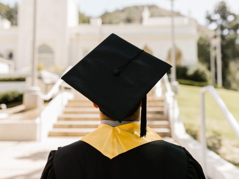 Back view of a graduate in a cap and gown standing outdoors facing a building.